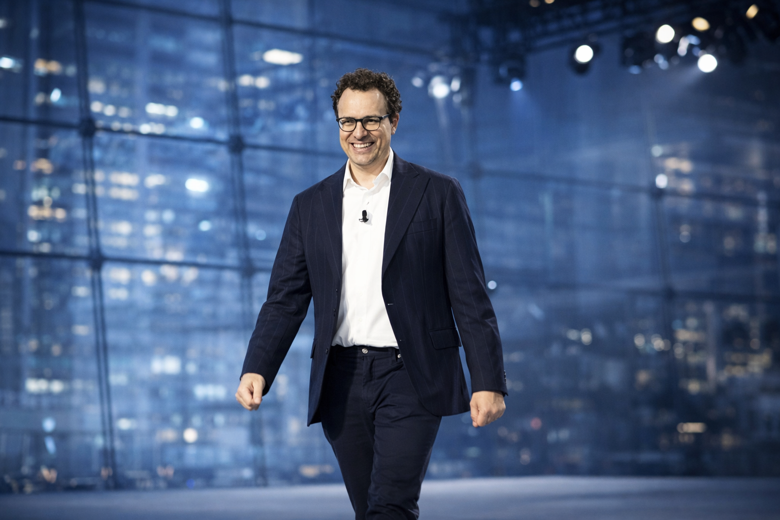 Dario Amodei entre en scène dans un cadre de conférence technologique, entre éclairages bleutés, fond architectural vitré et mise en scène sobre, cohérente avec l’univers de l’intelligence artificielle.