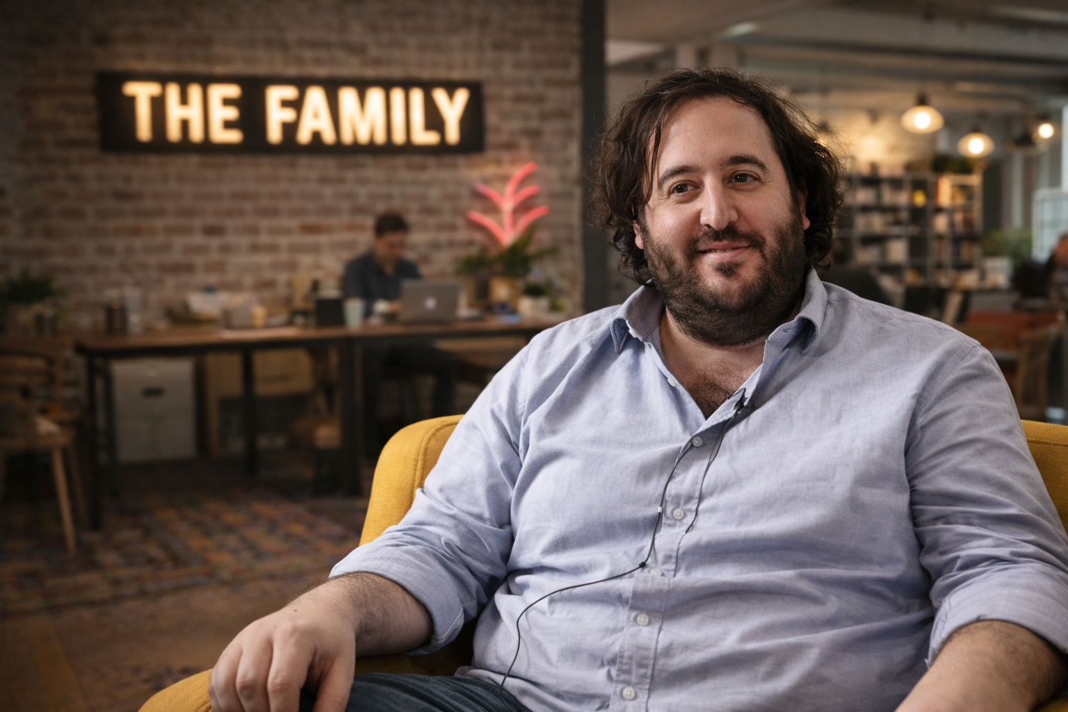 Portrait photographique en format paysage d’un homme assis dans un fauteuil jaune au sein d’un espace de coworking évoquant The Family, avec en arrière-plan un bureau animé, un mur en briques et une enseigne lumineuse « THE FAMILY », dans une ambiance professionnelle et startup.
