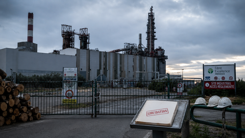 Vue éditoriale 16:9 de l’usine Fibre Excellence Provence à Tarascon, à l’arrêt derrière un portail fermé, avec des rondins de bois, des casques abandonnés et un dossier marqué « Liquidation ».