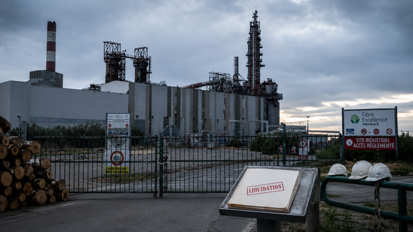 Vue éditoriale 16:9 de l’usine Fibre Excellence Provence à Tarascon, à l’arrêt derrière un portail fermé, avec des rondins de bois, des casques abandonnés et un dossier marqué « Liquidation ».