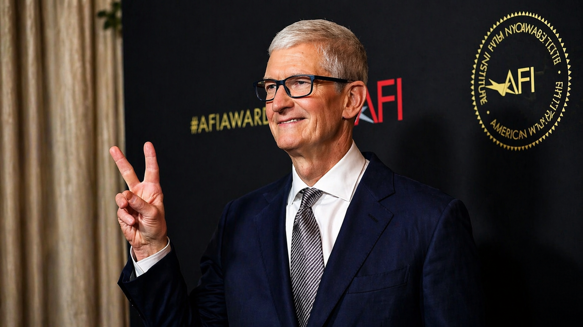 Portrait éditorial en format paysage de Tom Cook, vêtu d’un costume bleu marine et d’une cravate claire, levant un signe de victoire devant un fond de cérémonie noir marqué par des éléments visuels institutionnels. L’image évoque une prise de parole publique, une séquence de transition stratégique ou une annonce majeure au sommet d’un grand groupe technologique.
